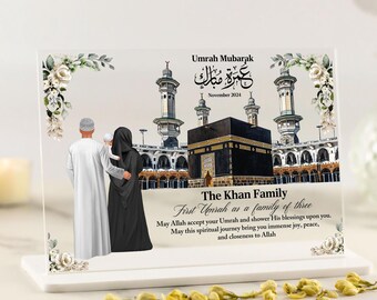 May include: A white acrylic plaque with a photo of the Kaaba in Mecca, Saudi Arabia. The plaque features a family of three, a man, a woman, and a child, standing in front of the Kaaba. The text on the plaque reads "Umrah Mubarak" in Arabic, "November 2024", "The Khan Family", "First Umrah as a family of three", "May Allah accept your Umrah and shower His blessings upon you. May this spiritual journey bring you immense joy, peace, and closeness to Allah."