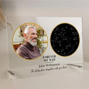 May include: A clear acrylic photo frame with a picture of a man in a brown turtleneck and a black and white star map. The text reads "FOREVER MY DAD" and "John Williamson". The frame is supported by two silver metal posts.