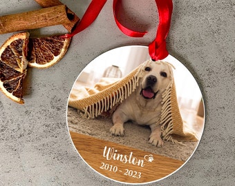May include: Round Christmas ornament featuring a photo of a dog under a blanket. The ornament has a wooden-look border with the name "Winston" and dates "2010-2023" printed in white. A red ribbon is attached for hanging.