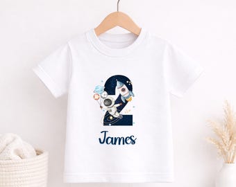 May include: White short-sleeved t-shirt featuring a colorful space-themed graphic. The design includes a number 2, an astronaut, a rocket ship, planets, and the name "James" in navy blue script. The shirt is hanging on a wooden hanger.