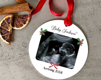 May include: A white, round Christmas ornament with a red ribbon. The ornament features a sonogram image, the text "Baby Jackson" and "Arriving 2024!" in black script, and holly accents. Cinnamon sticks and dried orange slices are in the background.