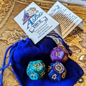 Puede incluir: Juego de dados Astro con una bolsa de terciopelo azul. Los dados son de color turquesa, morado y azul con símbolos y números dorados. El juego incluye dos tarjetas con el texto "Astro Dice" y "A Fun & Powerful Divination tool".