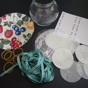 May include: A craft kit containing a clear glass jar, a circular fabric cover with a strawberry and berry print, a spool of red ribbon, a coil of green gingham ribbon, several yellow rubber bands, a stack of white circular paper discs, and a card.