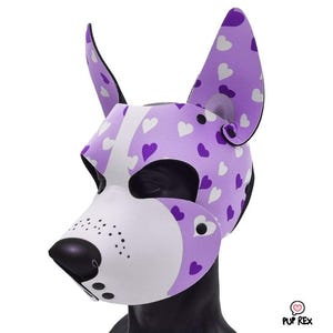 Puede incluir: Una máscara con forma de perro con hocico blanco y nariz negra. La máscara es principalmente lavanda con patrones de corazones blancos y morados. Las orejas están erguidas y también presentan el patrón de corazón. La máscara está diseñada para ser usada.