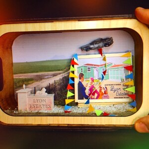 May include: A miniature diorama of a suburban neighbourhood with a small model car flying overhead. The diorama is set in a wooden frame and includes a sign that reads "Lyon Estates".
