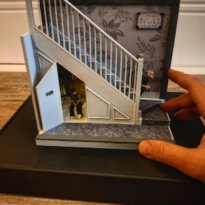 Może przedstawiać: Miniaturowy model schodów ze schowkiem pod schodami. Model przedstawia białe schody na tle szarej tapety w kwiaty. Znak głosi "Cupboard under the Stairs".