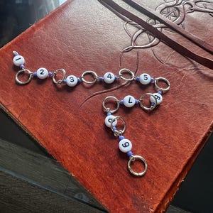 May include: Two silver-toned charm bracelets with white beads featuring black numbers and blue accents. The bracelets are on a brown leather-bound journal with Celtic knotwork. The numbers on the beads are 2, 3, 4, 5, 6, and L.