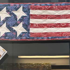 May include: A handmade American flag quilt with a blue and white star pattern on the left side and red and white stripes on the right side. The quilt is made with a bandana print fabric.