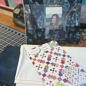 Może przedstawiać: Zestaw tkanin do patchworku z kwiatowym wzorem w odcieniach niebieskiego i białego. Zestaw jest zapakowany w przezroczystą plastikową torbę z etykietą z napisem "moda SHORELINE by Camille Roskelley". Zestaw zawiera również wzór patchworkowy zatytułowany "Nine Lives" z kolorowym wzorem patchworkowym.