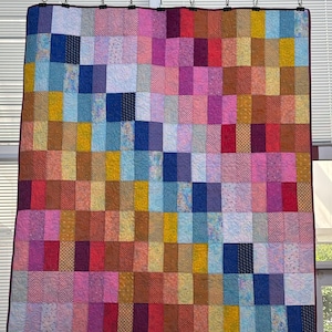 Könnte beinhalten: Ein rechteckiger Quilt mit einem Gitter aus bunten Stoffrechtecken in Blau-, Rosa-, Gelb-, Rot- und Brauntönen. Der Quilt hat einen dunkelvioletten Rand und ist durchgehend mit einem wirbelnden Muster abgesteppt.