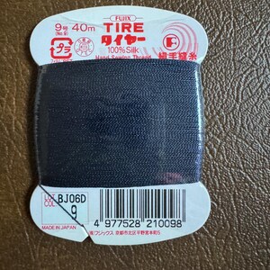 May include: A spool of black 100% silk thread for hand sewing. The spool is labeled "FUJIX TIRE ワイヤー 100% Silk Hand Sewing Thread 絹手縫糸" and has a barcode. The label also says "9号 40m (No.9) プラ" and "MADE IN JAPAN (株)フジックス 京都市北区平野宮本町5".