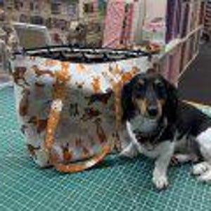Puede incluir: Una bolsa de tela blanca con estampado de dachshund marrón y naranja. La bolsa tiene asas naranjas y un marco de metal negro en la parte superior.