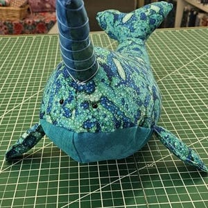 Puede incluir: Un juguete de peluche narval azul y verde con un cuerno enroscado. El juguete está hecho de tela con un patrón de burbujas y tiene ojos negros.
