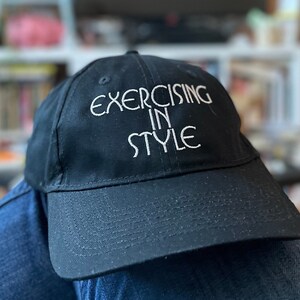 May include: Black baseball cap with the embroidered text "EXERCISING IN STYLE" in white. The cap has a curved brim and is made of a soft, durable fabric. A casual accessory for workouts or everyday wear.