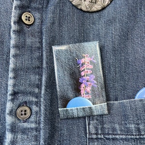 Peut inclure: Une chemise en jean avec une poche contenant un cadre en plastique transparent avec une fleur pressée à l'intérieur. La chemise a deux boutons et un bouton rond avec le texte "IT IS WHAT INSILVE" dessus.