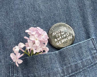 Könnte beinhalten: Ein schwarzer Knopf mit weißem Text, der "Another Jew Against Genocide" lautet, befindet sich in einer blauen Jeans-Tasche. Neben dem Knopf befindet sich ein Büschel hellrosa Blüten in der Tasche.