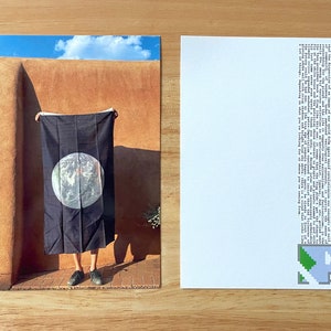 May include: A person holds a black flag with a white circle depicting the Earth in front of a tan adobe wall. The sky is blue with white clouds.