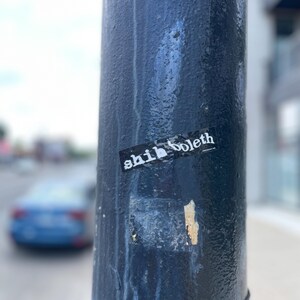 May include: A close-up of a dark blue painted pole with a small sticker that reads "shibboleth". The pole has streaks of paint and a weathered appearance. A blue car is blurred in the background.