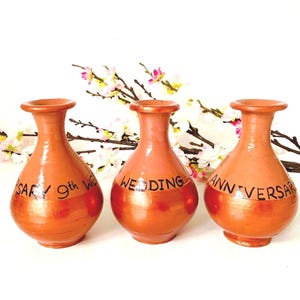 May include: Three terracotta-colored vases with a metallic gold band around the middle. The vases have text that reads "9th Wedding Anniversary" and "Wedding Anniversary". White and pink flowers are in the background.
