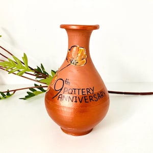 May include: Earthenware vase in a terracotta hue, featuring a painted floral motif and the inscription "9th Pottery Anniversary." The vase has a bulbous form and a slender neck. Greenery and flowers are in the background.
