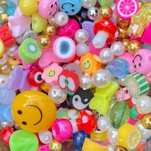 May include: A colorful assortment of plastic beads and charms, including smiley faces, hearts, stars, and fruit shapes. The beads are in various colors, including yellow, blue, green, red, and pink.