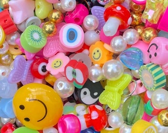 Peut inclure: Un assortiment coloré de perles et de breloques en plastique, comprenant des visages souriants, des cœurs, des étoiles et des formes de fruits. Les perles sont de différentes couleurs, notamment le jaune, le bleu, le vert, le rouge et le rose.
