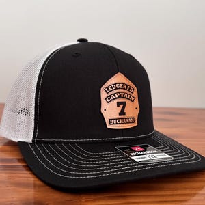 May include: Black and white trucker hat with a curved brim. The front panel is black with a leather patch that reads "LEDGERTO CAPTAIN 7 BUCHANAN". The back is white mesh. The hat is sitting on a wooden surface.