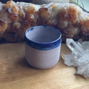 May include: A small, speckled ceramic cup with a blue rim. The cup is a light pink color and sits on a wooden surface. Crystals and a large cluster of orange-yellow stones are in the background.