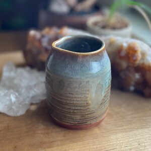 May include: A handmade ceramic vase with a textured, ribbed design. The vase features a color gradient, transitioning from a reddish-brown base to a light blue-green midsection, and a brown rim. The interior is dark. The vase is on a wooden surface.