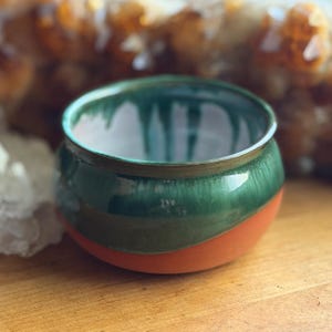 May include: A small, round ceramic bowl featuring a smooth, glossy dark green glaze that drips down from the rim over a matte, unglazed terracotta-colored base. The interior of the bowl is finished with a white glaze, creating a contrast.