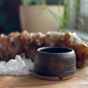 May include: A small, dark charcoal-colored ceramic bowl with a textured, horizontal-lined exterior and a smooth, light-tan unglazed base. The bowl features a subtle, vertical notch at the bottom edge, contrasting with its dark, matte finish.