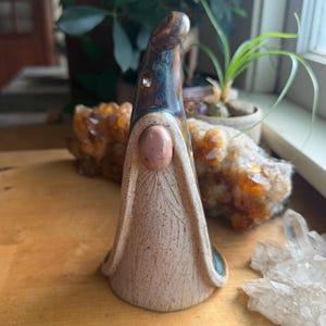 May include: A ceramic gnome figurine with a tall, dark brown glazed hat featuring a small embedded crystal. The body is a light beige, speckled clay with a smooth, rounded pink nose and carved vertical lines resembling a long, flowing beard.