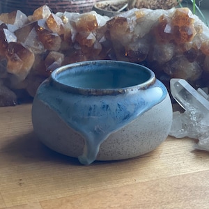 May include: A small, round ceramic bowl with a speckled, off-white base and a light blue glaze that drips down the side. The rim has a brown accent. The bowl is likely handmade and suitable for holding small items.