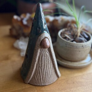 May include: A ceramic gnome figurine with a pointed teal hat and a textured, tan beard. The gnome has a small, rounded nose and is set against a blurred background with plants. The figurine is handmade and has a rustic appearance.