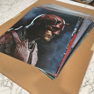May include: A collection of art prints depicting a comic book character in a red and black costume, with a horned mask. The prints are in protective sleeves and are on a brown cardboard backing.