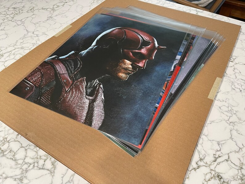 May include: A collection of art prints depicting a comic book character in a red and black costume, with a horned mask. The prints are in protective sleeves and are on a brown cardboard backing.