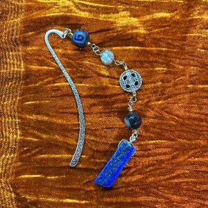 May include: A silver bookmark with a decorative swirl design. The bookmark has a chain with various beads, including a blue and white bead, a white bead, a black and white bead, and a blue stone.