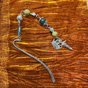 May include: A silver bookmark with a decorative design and a charm featuring a crown and a sword. The charm is attached to a chain with green and brown beads.
