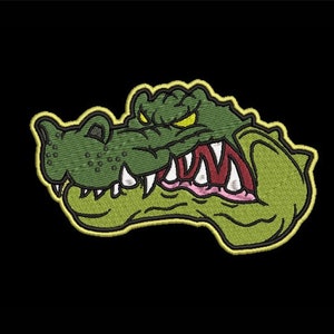 Può includere: Testa di alligatore verde con occhi gialli e denti affilati, ricamata su una toppa con un contorno giallo.