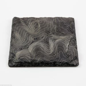 May include: Square, dark grey slate coaster featuring a white topographic map design. The coaster has a natural, rough edge and a detailed, swirling pattern. The design resembles contour lines, creating a textured, artistic appearance. A decorative item for the home.