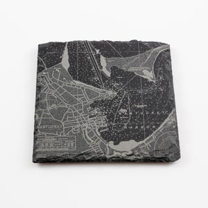 May include: A square, dark gray slate coaster featuring a detailed map of Nantucket Island. The map is etched in a lighter gray, showing streets, water, and the words "Nantucket Harbor". The coaster has a natural, textured edge.