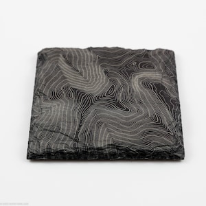 May include: Square, dark gray coaster with a topographic map design. The coaster features a detailed pattern of contour lines in a lighter gray color, creating a visual representation of elevation changes. The edges of the coaster have a slightly rough texture.
