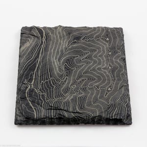 May include: A square, dark gray slate coaster with a topographic map design etched into the surface. The map features contour lines and a dotted path with triangle markers, creating a textured, natural aesthetic.