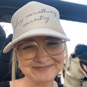 Girl wearing tan “Play Something Country” foam trucker hat in an offroad vehicle with a dog in the background, Western cowgirl style.