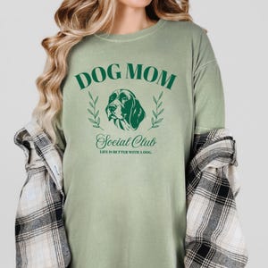 May include: Green long-sleeved t-shirt with the text "Dog Mom Social Club" and a graphic of a dog's head. The text below the graphic reads "Life is better with a dog."
