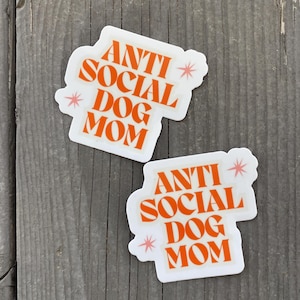 May include: Two white stickers with orange text that reads "Anti Social Dog Mom" with pink stars on a wood background.