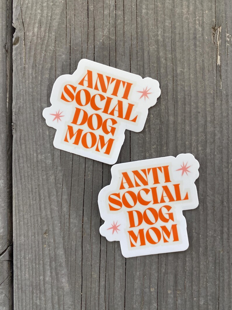 May include: Two white stickers with orange text that reads "Anti Social Dog Mom" with pink stars on a wood background.