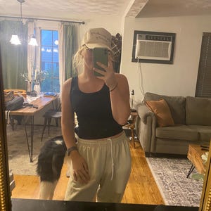 Girl taking mirror selfie wearing tan “Play Something Country” trucker hat, hair up, black tank top, and tan sweatpants, gold mirror and dog in dining room background.