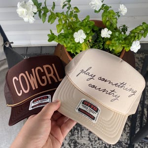 Hand holding tan “Play Something Country” trucker hat next to brown “COWGRL” hat with tan lettering, porch setting with pot of white flowers.