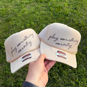 Hand holding two tan “Play Something Country” Otto high front foam trucker hat with dark brown lettering in front of green grass, retro Western cowgirl style cap with slim custom gold hat chain.